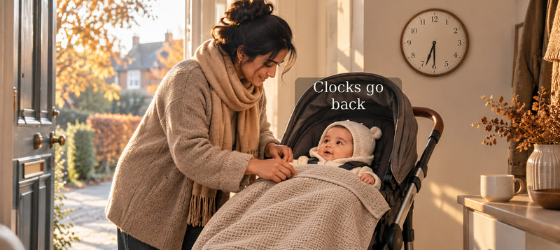 Parent preparing a baby in a pram by an autumn doorway, with a coffee-coloured cellular cotton blanket and a clock showing 6:30. Text reads “Clocks go back.”