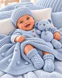 Blue 9XB720-B knitted cotton baby gift set in use: smiling baby wearing blue knitted beanie and booties beside matching teddy and blanket